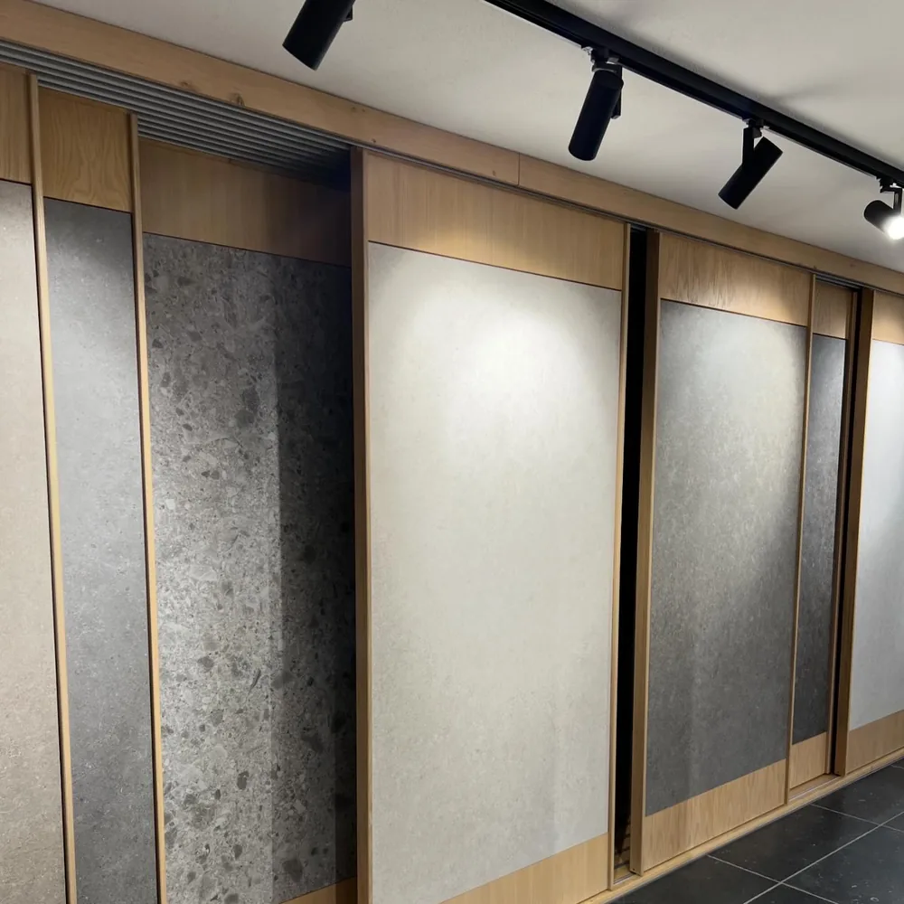 Musterregal im Showroom mit großformatigen Platten in Hellgrau, Anthrazit und Terrazzo, in hellen Holzrahmen auf Schienen, darunter dunkler Boden und Strahler an der Decke
