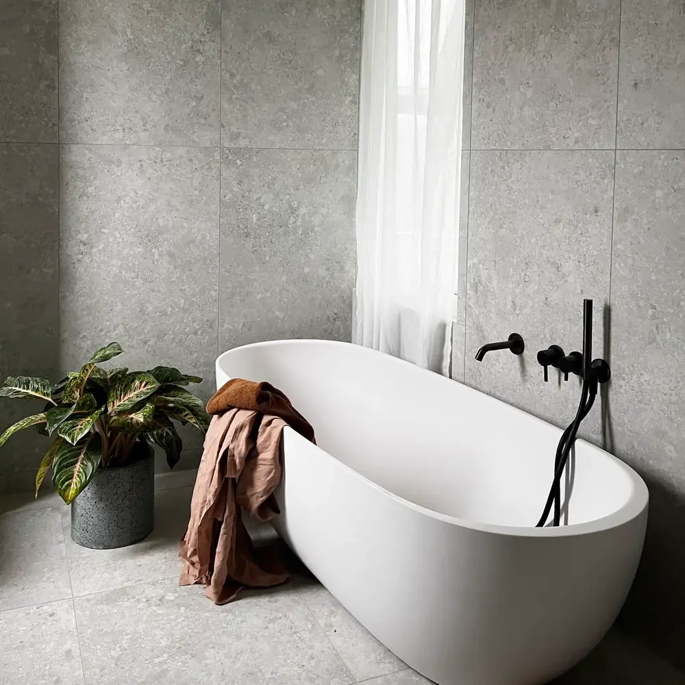 Bad mit grauen Natursteinfliesen, freistehender Badewanne mit schwarzer Wandarmatur und grüner Pflanze